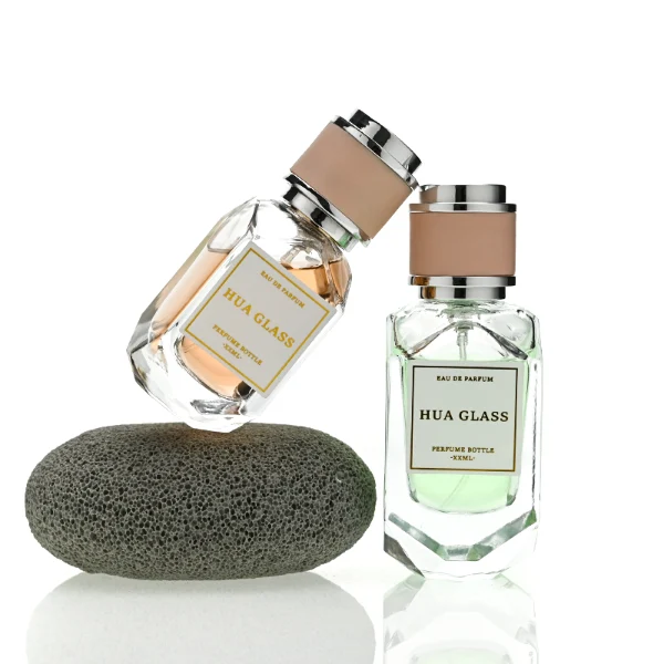 Two multi-faceted, gemstone-shaped clear glass perfume bottles with thick faceted bases, holding peach and light green liquids. Each bottle features a square white label and is topped with a unique cylindrical cap showcasing a matte beige center band paired with polished silver metallic trim.