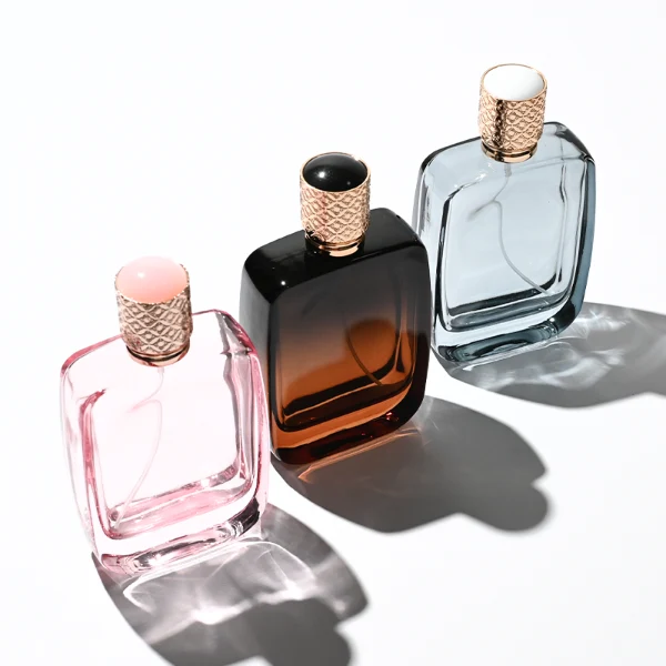 Three flat rectangular perfume bottles with rounded corners are shown in a row. The bottles feature semi-translucent glass bodies in soft grey-blue, delicate pink, and a dark amber-to-black gradient. Each is paired with a cylindrical rose-gold cap detailed with an intricate engraved geometric pattern and capped with a smooth, domed insert on top.