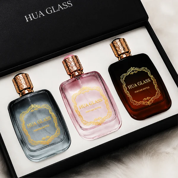 Three flat rectangular perfume bottles with rounded corners are shown in a row. The bottles feature semi-translucent glass bodies in soft grey-blue, delicate pink, and a dark amber-to-black gradient. Each is paired with a cylindrical rose-gold cap detailed with an intricate engraved geometric pattern and capped with a smooth, domed insert on top.