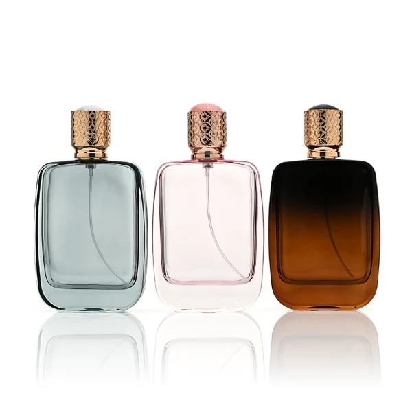 Three flat rectangular perfume bottles with rounded corners are shown in a row. The bottles feature semi-translucent glass bodies in soft grey-blue, delicate pink, and a dark amber-to-black gradient. Each is paired with a cylindrical rose-gold cap detailed with an intricate engraved geometric pattern and capped with a smooth, domed insert on top.