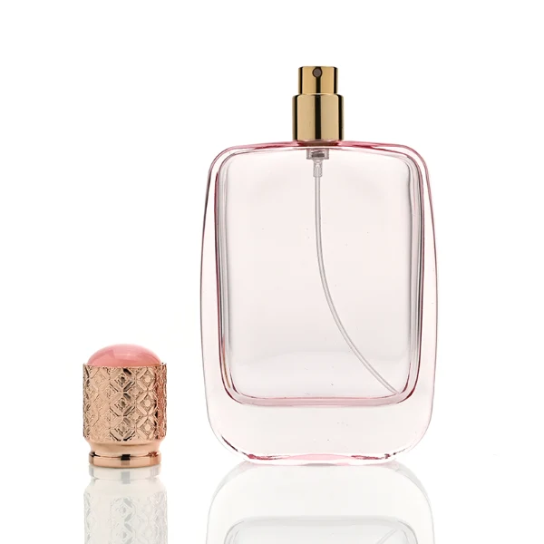 Three flat rectangular perfume bottles with rounded corners are shown in a row. The bottles feature semi-translucent glass bodies in soft grey-blue, delicate pink, and a dark amber-to-black gradient. Each is paired with a cylindrical rose-gold cap detailed with an intricate engraved geometric pattern and capped with a smooth, domed insert on top.