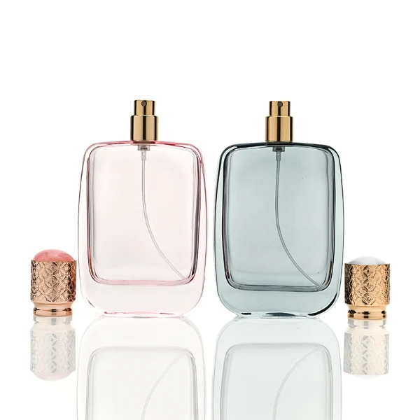 Three flat rectangular perfume bottles with rounded corners are shown in a row. The bottles feature semi-translucent glass bodies in soft grey-blue, delicate pink, and a dark amber-to-black gradient. Each is paired with a cylindrical rose-gold cap detailed with an intricate engraved geometric pattern and capped with a smooth, domed insert on top.