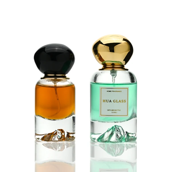 Two cylindrical clear glass perfume bottles filled with amber and light green liquids, featuring a unique, thick base internally sculpted into a mountain peak design. The bottles are paired with smooth, oversized pebble-shaped bulbous caps shown in glossy black and reflective polished gold finishes.