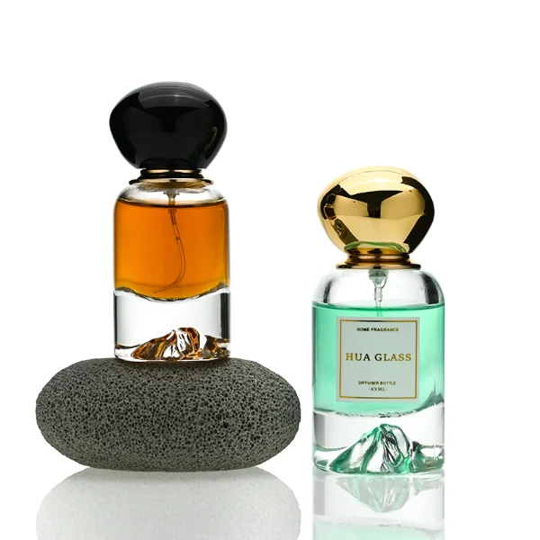 Two cylindrical clear glass perfume bottles filled with amber and light green liquids, featuring a unique, thick base internally sculpted into a mountain peak design. The bottles are paired with smooth, oversized pebble-shaped bulbous caps shown in glossy black and reflective polished gold finishes.