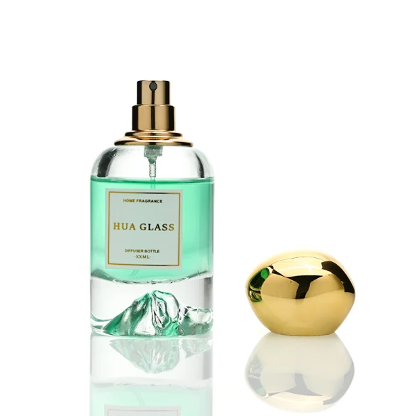 Two cylindrical clear glass perfume bottles filled with amber and light green liquids, featuring a unique, thick base internally sculpted into a mountain peak design. The bottles are paired with smooth, oversized pebble-shaped bulbous caps shown in glossy black and reflective polished gold finishes.