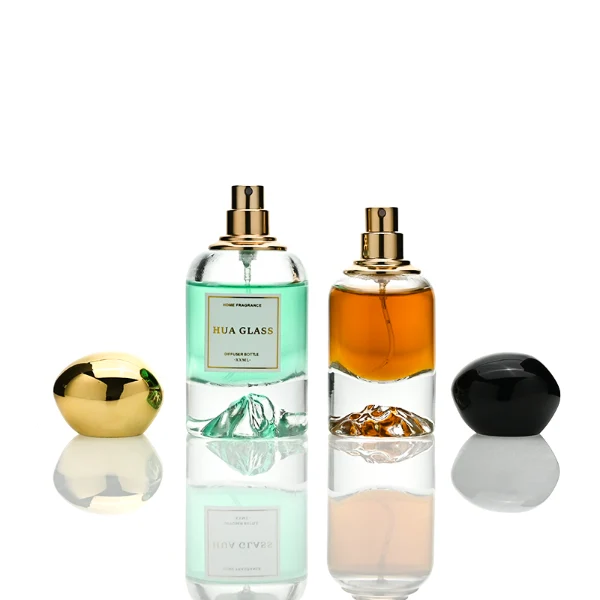 Two cylindrical clear glass perfume bottles filled with amber and light green liquids, featuring a unique, thick base internally sculpted into a mountain peak design. The bottles are paired with smooth, oversized pebble-shaped bulbous caps shown in glossy black and reflective polished gold finishes.