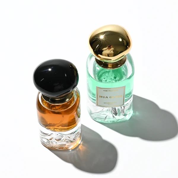 Two cylindrical clear glass perfume bottles filled with amber and light green liquids, featuring a unique, thick base internally sculpted into a mountain peak design. The bottles are paired with smooth, oversized pebble-shaped bulbous caps shown in glossy black and reflective polished gold finishes.