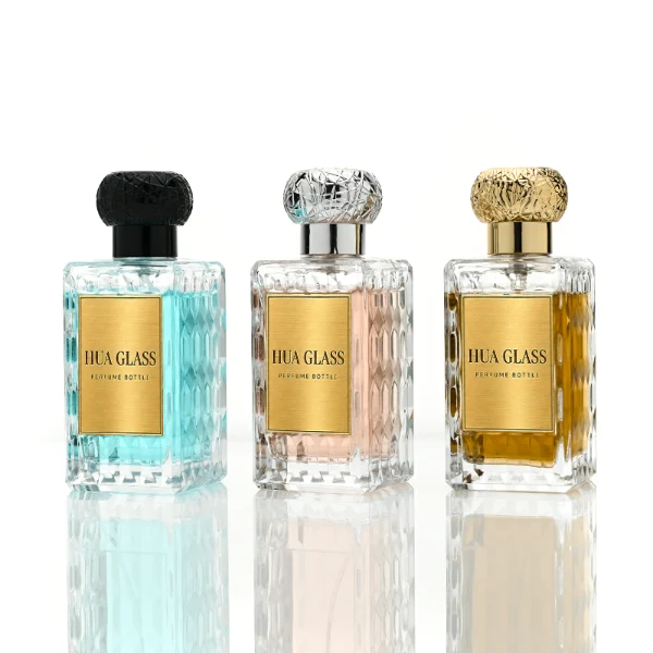 three clear rectangular glass perfume bottles feature deeply textured, diamond-faceted faces and geometric patterned side walls, holding bright blue, pale pink, and deep amber liquids. Each bottle is topped with an ornate, spherical textured ball cap featuring a heavily cracked or webbed aesthetic, shown in matte black, polished silver, and bright gold finishes.