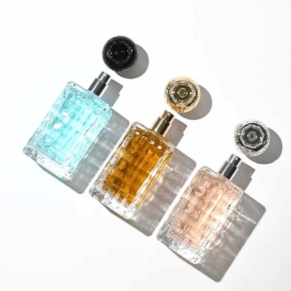three clear rectangular glass perfume bottles feature deeply textured, diamond-faceted faces and geometric patterned side walls, holding bright blue, pale pink, and deep amber liquids. Each bottle is topped with an ornate, spherical textured ball cap featuring a heavily cracked or webbed aesthetic, shown in matte black, polished silver, and bright gold finishes.