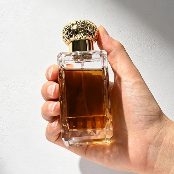 three clear rectangular glass perfume bottles feature deeply textured, diamond-faceted faces and geometric patterned side walls, holding bright blue, pale pink, and deep amber liquids. Each bottle is topped with an ornate, spherical textured ball cap featuring a heavily cracked or webbed aesthetic, shown in matte black, polished silver, and bright gold finishes.