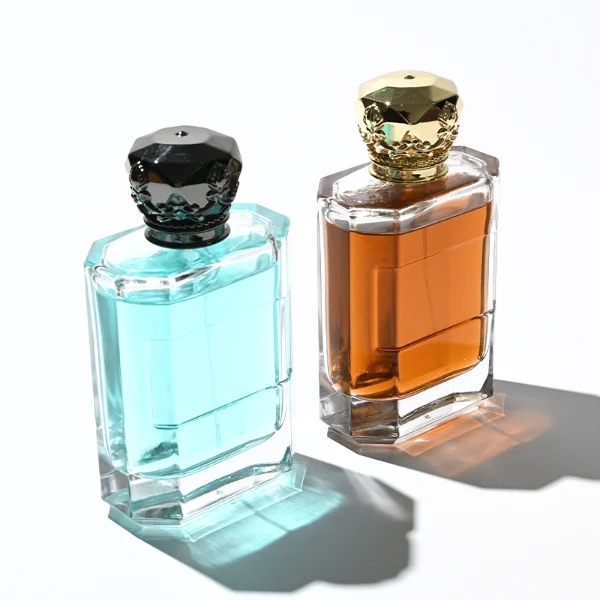 Two flat rectangular clear glass perfume bottles are shown with faceted, angled bottom corners and thick glass bases, holding light blue and amber liquids. Each bottle features a subtle debossed rectangular label panel on the front and is topped with an ornate, heavy Zamac crown-motif cap with a faceted dome, finished in glossy black and polished gold.