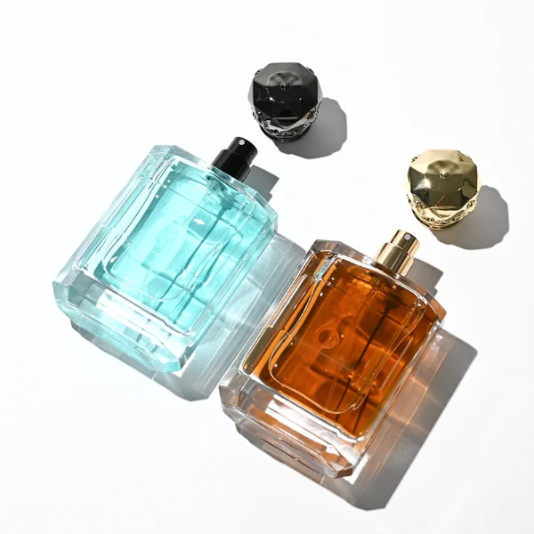 Two flat rectangular clear glass perfume bottles are shown with faceted, angled bottom corners and thick glass bases, holding light blue and amber liquids. Each bottle features a subtle debossed rectangular label panel on the front and is topped with an ornate, heavy Zamac crown-motif cap with a faceted dome, finished in glossy black and polished gold.