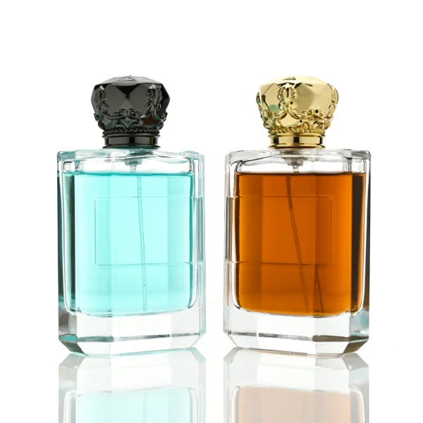 Two flat rectangular clear glass perfume bottles are shown with faceted, angled bottom corners and thick glass bases, holding light blue and amber liquids. Each bottle features a subtle debossed rectangular label panel on the front and is topped with an ornate, heavy Zamac crown-motif cap with a faceted dome, finished in glossy black and polished gold.