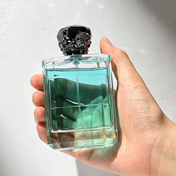Two flat rectangular clear glass perfume bottles are shown with faceted, angled bottom corners and thick glass bases, holding light blue and amber liquids. Each bottle features a subtle debossed rectangular label panel on the front and is topped with an ornate, heavy Zamac crown-motif cap with a faceted dome, finished in glossy black and polished gold.
