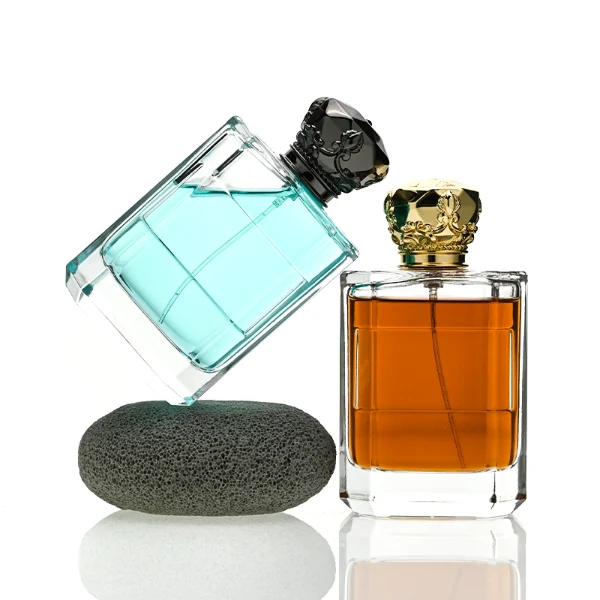 Two flat rectangular clear glass perfume bottles are shown with faceted, angled bottom corners and thick glass bases, holding light blue and amber liquids. Each bottle features a subtle debossed rectangular label panel on the front and is topped with an ornate, heavy Zamac crown-motif cap with a faceted dome, finished in glossy black and polished gold.
