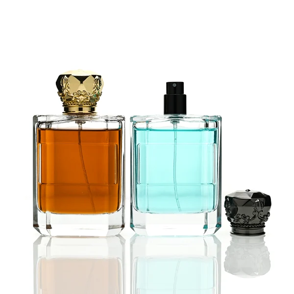 Two flat rectangular clear glass perfume bottles are shown with faceted, angled bottom corners and thick glass bases, holding light blue and amber liquids. Each bottle features a subtle debossed rectangular label panel on the front and is topped with an ornate, heavy Zamac crown-motif cap with a faceted dome, finished in glossy black and polished gold.