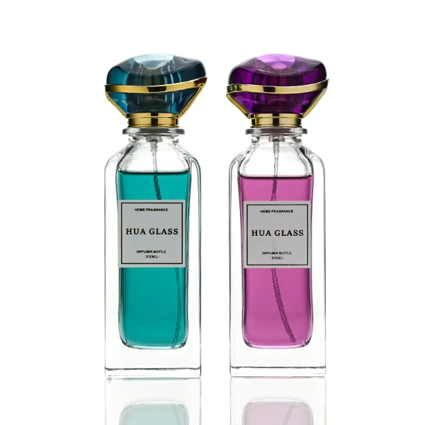 two tall rectangular clear glass perfume bottles with thick bases and curved shoulders are shown holding bright teal and magenta liquids. Each bottle features a white rectangular front label and is topped with an oversized, translucent jewel-shaped cap split by a metallic gold trim band, presented in matching teal and purple colors.