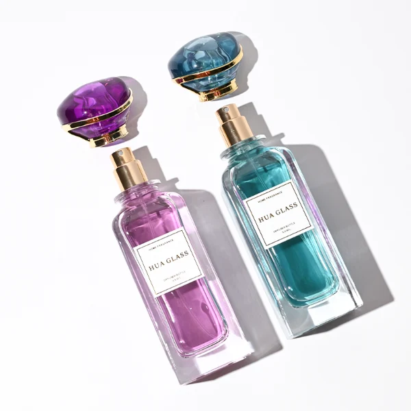 two tall rectangular clear glass perfume bottles with thick bases and curved shoulders are shown holding bright teal and magenta liquids. Each bottle features a white rectangular front label and is topped with an oversized, translucent jewel-shaped cap split by a metallic gold trim band, presented in matching teal and purple colors.