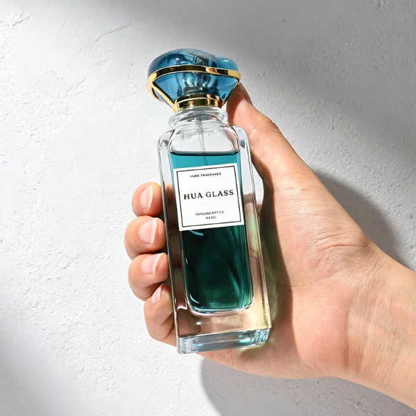two tall rectangular clear glass perfume bottles with thick bases and curved shoulders are shown holding bright teal and magenta liquids. Each bottle features a white rectangular front label and is topped with an oversized, translucent jewel-shaped cap split by a metallic gold trim band, presented in matching teal and purple colors.