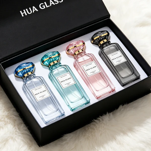 two tall rectangular clear glass perfume bottles with thick bases and curved shoulders are shown holding bright teal and magenta liquids. Each bottle features a white rectangular front label and is topped with an oversized, translucent jewel-shaped cap split by a metallic gold trim band, presented in matching teal and purple colors.