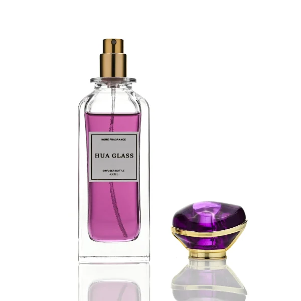 two tall rectangular clear glass perfume bottles with thick bases and curved shoulders are shown holding bright teal and magenta liquids. Each bottle features a white rectangular front label and is topped with an oversized, translucent jewel-shaped cap split by a metallic gold trim band, presented in matching teal and purple colors.
