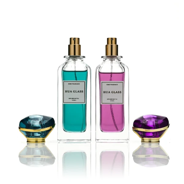 two tall rectangular clear glass perfume bottles with thick bases and curved shoulders are shown holding bright teal and magenta liquids. Each bottle features a white rectangular front label and is topped with an oversized, translucent jewel-shaped cap split by a metallic gold trim band, presented in matching teal and purple colors.