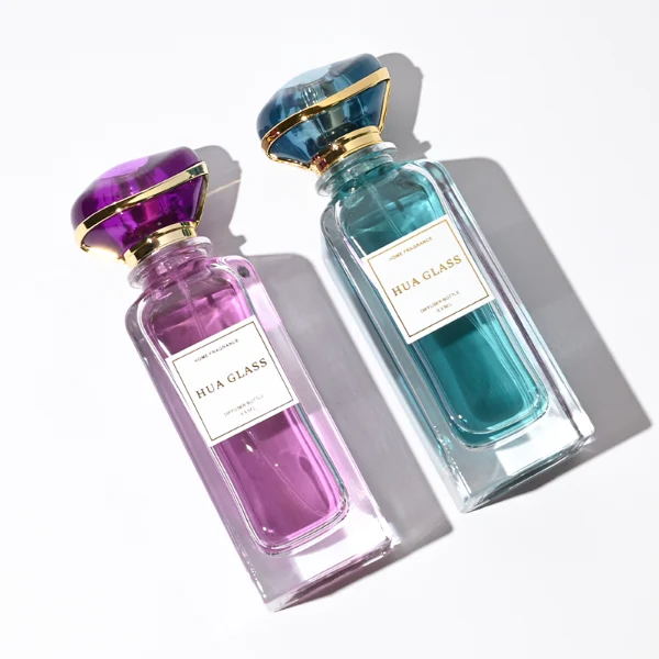 two tall rectangular clear glass perfume bottles with thick bases and curved shoulders are shown holding bright teal and magenta liquids. Each bottle features a white rectangular front label and is topped with an oversized, translucent jewel-shaped cap split by a metallic gold trim band, presented in matching teal and purple colors.