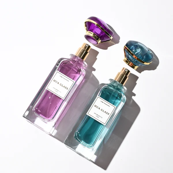 two tall rectangular clear glass perfume bottles with thick bases and curved shoulders are shown holding bright teal and magenta liquids. Each bottle features a white rectangular front label and is topped with an oversized, translucent jewel-shaped cap split by a metallic gold trim band, presented in matching teal and purple colors.