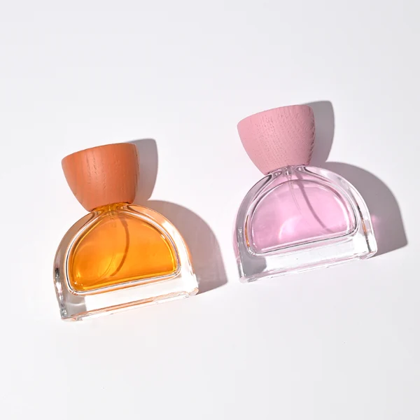 three clear glass perfume bottles feature a distinctive semi-circular arch silhouette with a flat base, holding vibrant orange, soft pink, and bright yellow liquids. Each bottle is topped with an upward-flaring, wide-mouth natural solid wood cap displaying prominent wood-grain textures, finished in stained shades of terracotta orange, dusty rose pink, and mustard yellow.