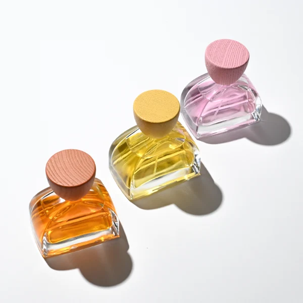 three clear glass perfume bottles feature a distinctive semi-circular arch silhouette with a flat base, holding vibrant orange, soft pink, and bright yellow liquids. Each bottle is topped with an upward-flaring, wide-mouth natural solid wood cap displaying prominent wood-grain textures, finished in stained shades of terracotta orange, dusty rose pink, and mustard yellow.