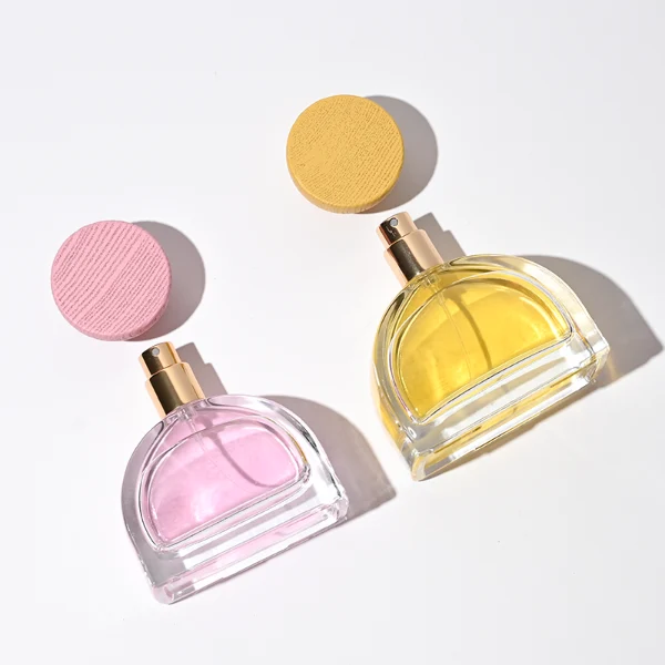 three clear glass perfume bottles feature a distinctive semi-circular arch silhouette with a flat base, holding vibrant orange, soft pink, and bright yellow liquids. Each bottle is topped with an upward-flaring, wide-mouth natural solid wood cap displaying prominent wood-grain textures, finished in stained shades of terracotta orange, dusty rose pink, and mustard yellow.