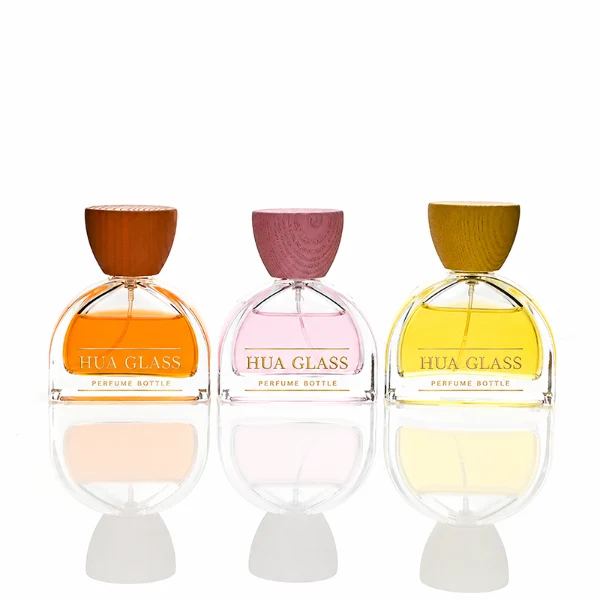 three clear glass perfume bottles feature a distinctive semi-circular arch silhouette with a flat base, holding vibrant orange, soft pink, and bright yellow liquids. Each bottle is topped with an upward-flaring, wide-mouth natural solid wood cap displaying prominent wood-grain textures, finished in stained shades of terracotta orange, dusty rose pink, and mustard yellow.