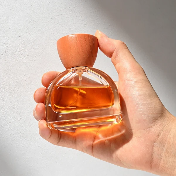 three clear glass perfume bottles feature a distinctive semi-circular arch silhouette with a flat base, holding vibrant orange, soft pink, and bright yellow liquids. Each bottle is topped with an upward-flaring, wide-mouth natural solid wood cap displaying prominent wood-grain textures, finished in stained shades of terracotta orange, dusty rose pink, and mustard yellow.