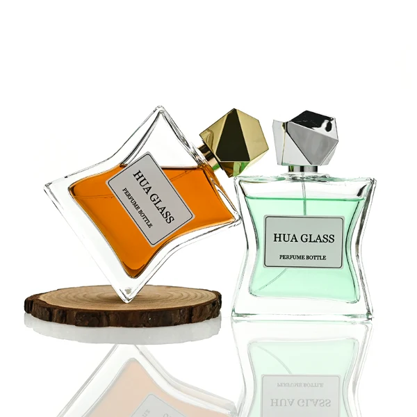 Two clear rectangular glass perfume bottles with a distinctively sculpted waisted design holding amber and light green liquids are shown. Each bottle features a central square white label and is topped with an asymmetrical, irregular multifaceted geometric cap featuring sharp reflective angles, presented in polished gold and high-shine chrome silver finishes.