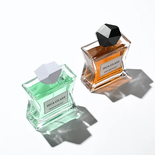 Two clear rectangular glass perfume bottles with a distinctively sculpted waisted design holding amber and light green liquids are shown. Each bottle features a central square white label and is topped with an asymmetrical, irregular multifaceted geometric cap featuring sharp reflective angles, presented in polished gold and high-shine chrome silver finishes.