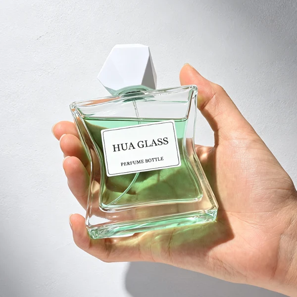 Two clear rectangular glass perfume bottles with a distinctively sculpted waisted design holding amber and light green liquids are shown. Each bottle features a central square white label and is topped with an asymmetrical, irregular multifaceted geometric cap featuring sharp reflective angles, presented in polished gold and high-shine chrome silver finishes.