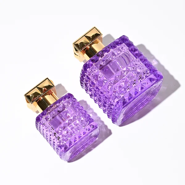 Two square semi-translucent violet glass perfume bottles are shown in 50ml and 100ml sizes, featuring an all-over rockstud-inspired studded diamond texture surrounding a smooth rectangular center panel. Each bottle is topped with a heavy, premium polished golden Zamac square geometric cap with a short cylindrical collar.