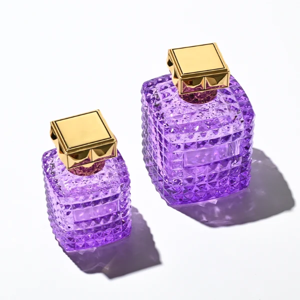 Two square semi-translucent violet glass perfume bottles are shown in 50ml and 100ml sizes, featuring an all-over rockstud-inspired studded diamond texture surrounding a smooth rectangular center panel. Each bottle is topped with a heavy, premium polished golden Zamac square geometric cap with a short cylindrical collar.