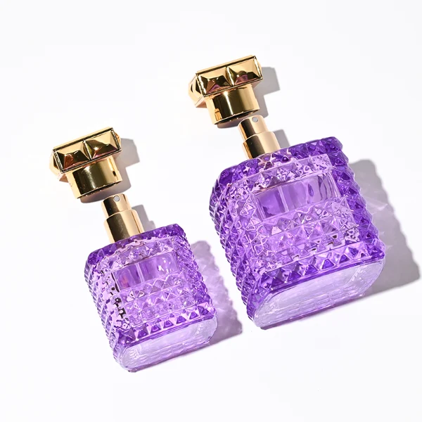 Two square semi-translucent violet glass perfume bottles are shown in 50ml and 100ml sizes, featuring an all-over rockstud-inspired studded diamond texture surrounding a smooth rectangular center panel. Each bottle is topped with a heavy, premium polished golden Zamac square geometric cap with a short cylindrical collar.