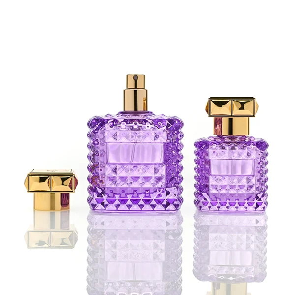 Two square semi-translucent violet glass perfume bottles are shown in 50ml and 100ml sizes, featuring an all-over rockstud-inspired studded diamond texture surrounding a smooth rectangular center panel. Each bottle is topped with a heavy, premium polished golden Zamac square geometric cap with a short cylindrical collar.