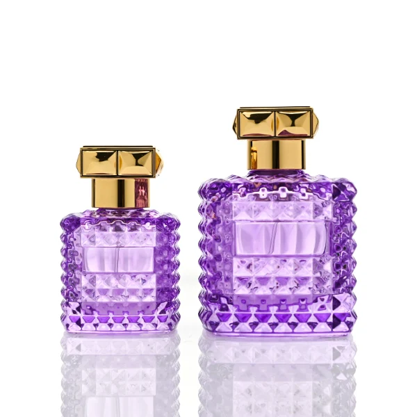 Two square semi-translucent violet glass perfume bottles are shown in 50ml and 100ml sizes, featuring an all-over rockstud-inspired studded diamond texture surrounding a smooth rectangular center panel. Each bottle is topped with a heavy, premium polished golden Zamac square geometric cap with a short cylindrical collar.