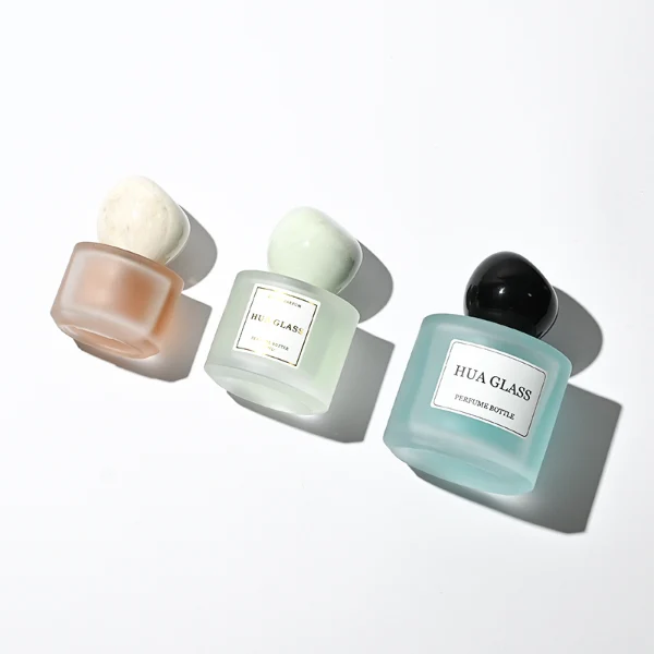 Three frosted semi-translucent cylindrical glass perfume bottles are shown in varying heights, containing amber, very light green, and sky blue liquids. The middle and right bottles are adorned with square white brand labels. Each bottle is topped with an oversized, asymmetrical stone-like pebble cap presented in a cream marble swirl, a pale jade green marble swirl, and a solid glossy black finish.