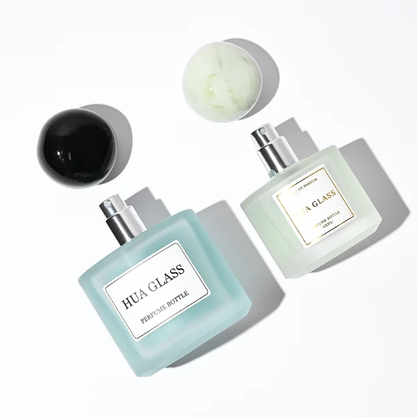 Three frosted semi-translucent cylindrical glass perfume bottles are shown in varying heights, containing amber, very light green, and sky blue liquids. The middle and right bottles are adorned with square white brand labels. Each bottle is topped with an oversized, asymmetrical stone-like pebble cap presented in a cream marble swirl, a pale jade green marble swirl, and a solid glossy black finish.