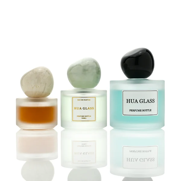 Three frosted semi-translucent cylindrical glass perfume bottles are shown in varying heights, containing amber, very light green, and sky blue liquids. The middle and right bottles are adorned with square white brand labels. Each bottle is topped with an oversized, asymmetrical stone-like pebble cap presented in a cream marble swirl, a pale jade green marble swirl, and a solid glossy black finish.