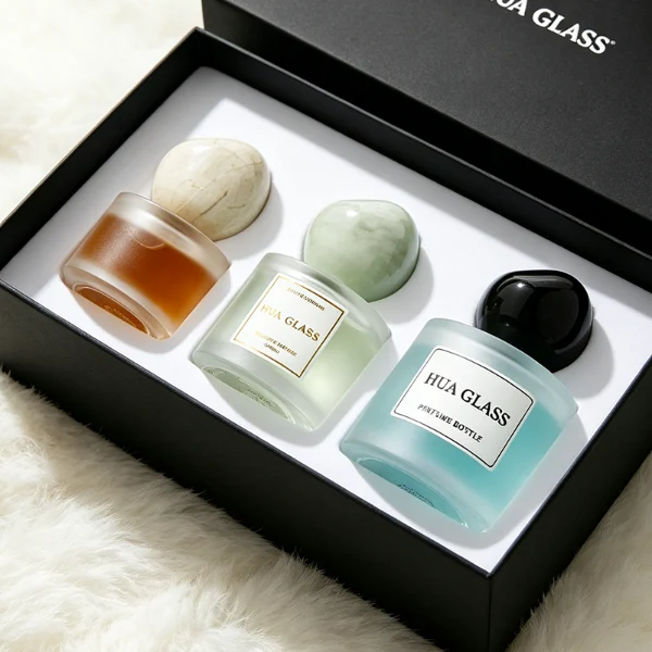 Three frosted semi-translucent cylindrical glass perfume bottles are shown in varying heights, containing amber, very light green, and sky blue liquids. The middle and right bottles are adorned with square white brand labels. Each bottle is topped with an oversized, asymmetrical stone-like pebble cap presented in a cream marble swirl, a pale jade green marble swirl, and a solid glossy black finish.