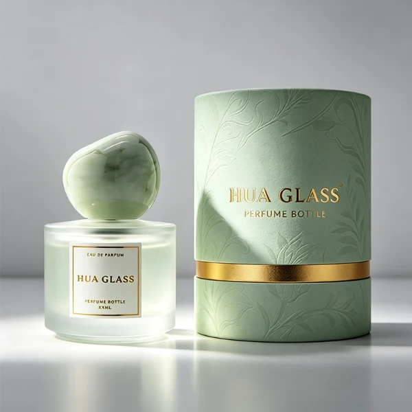 Three frosted semi-translucent cylindrical glass perfume bottles are shown in varying heights, containing amber, very light green, and sky blue liquids. The middle and right bottles are adorned with square white brand labels. Each bottle is topped with an oversized, asymmetrical stone-like pebble cap presented in a cream marble swirl, a pale jade green marble swirl, and a solid glossy black finish.
