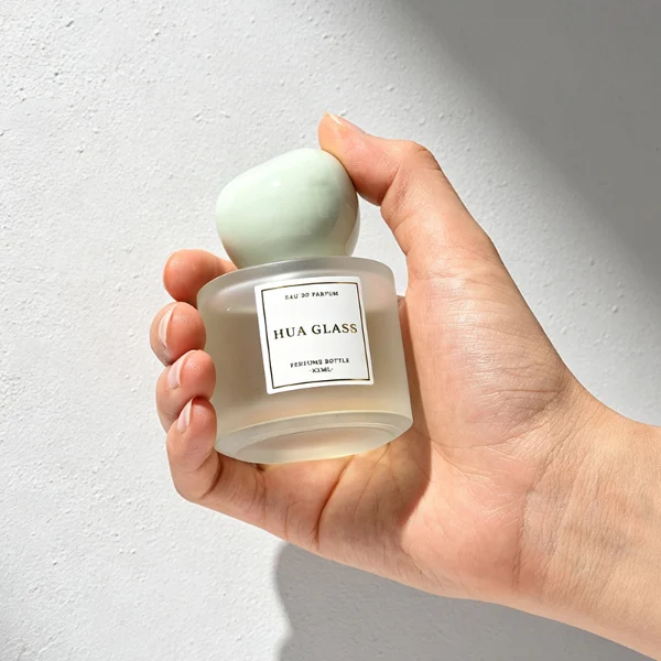 Three frosted semi-translucent cylindrical glass perfume bottles are shown in varying heights, containing amber, very light green, and sky blue liquids. The middle and right bottles are adorned with square white brand labels. Each bottle is topped with an oversized, asymmetrical stone-like pebble cap presented in a cream marble swirl, a pale jade green marble swirl, and a solid glossy black finish.