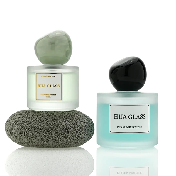Three frosted semi-translucent cylindrical glass perfume bottles are shown in varying heights, containing amber, very light green, and sky blue liquids. The middle and right bottles are adorned with square white brand labels. Each bottle is topped with an oversized, asymmetrical stone-like pebble cap presented in a cream marble swirl, a pale jade green marble swirl, and a solid glossy black finish.