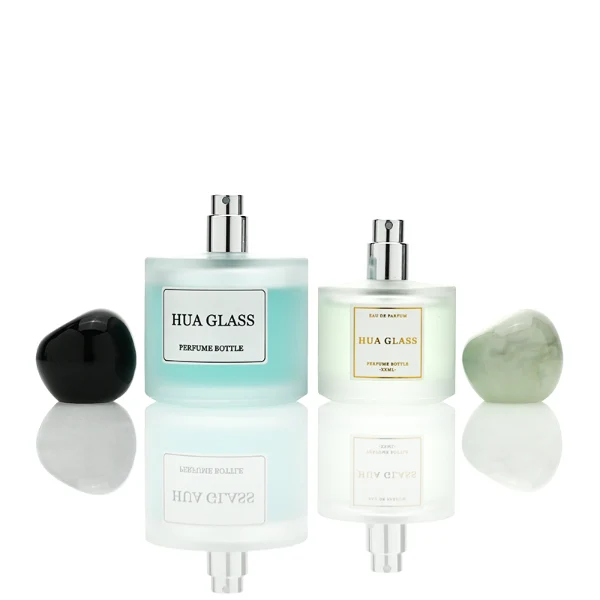 Three frosted semi-translucent cylindrical glass perfume bottles are shown in varying heights, containing amber, very light green, and sky blue liquids. The middle and right bottles are adorned with square white brand labels. Each bottle is topped with an oversized, asymmetrical stone-like pebble cap presented in a cream marble swirl, a pale jade green marble swirl, and a solid glossy black finish.