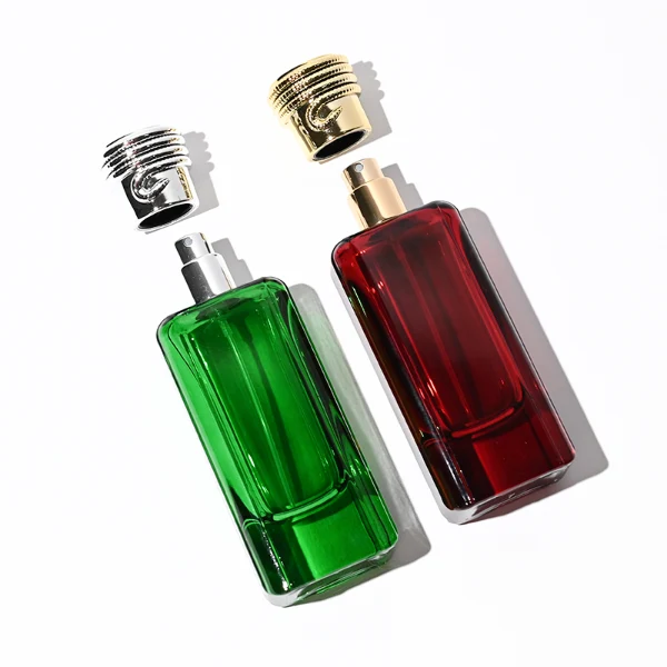 two tall rectangular, thick-bottomed glass perfume bottles are shown standing side by side. One bottle is made of vivid semi-translucent red glass and the other is vibrant semi-translucent green glass. Each bottle features subtly debossed vertical panels along its sides and is topped with a cylindrical cap encircled by a textured, coiling snake pattern—presented in polished gold on the red bottle and polished silver on the green bottle.